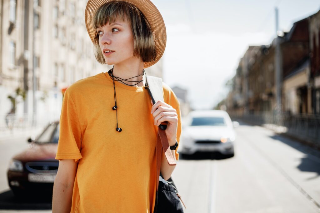 Young girl with headphones in a yellow T-shirt and a straw hat walks with a backpack along a city
